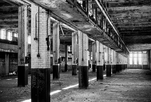 Black and white image of an abandoned industrial space with tall columns and scattered debris on the floor.