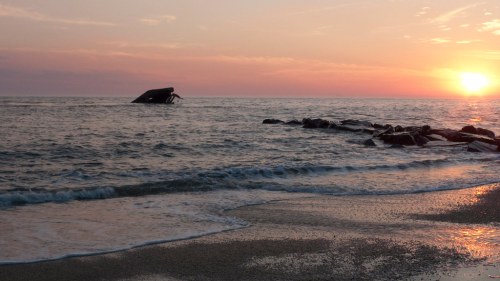A serene beach at sunset, with gentle waves and a shipwreck partially submerged in the water.