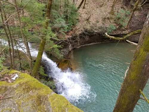 A serene waterfall cascading into a clear blue pool, surrounded by lush green trees and rocky terrain.