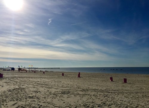 A serene beach scene with a clear sky, gentle waves, and colorful flags near a pier in the distance.