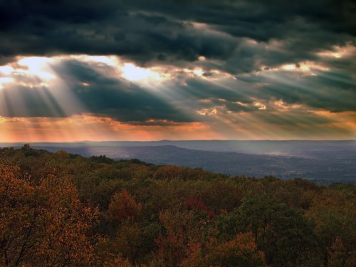 Dramatic sky with rays of sunlight breaking through clouds over a colorful autumn landscape.