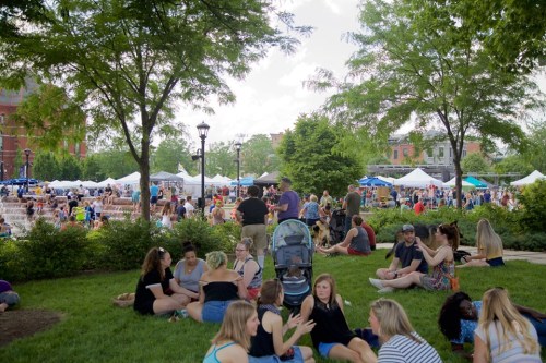 A lively outdoor event with people sitting on grass, food stalls, and tents in the background under a sunny sky.