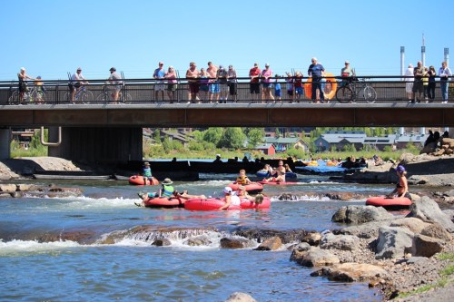 People float on inner tubes in a river while spectators watch from a bridge above on a sunny day.