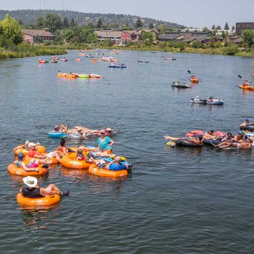 People floating on colorful inner tubes in a river, enjoying a sunny day with trees and buildings in the background.