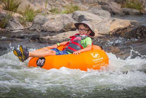 A child in a hat relaxes on an orange inner tube, floating down a rocky river with splashing water.