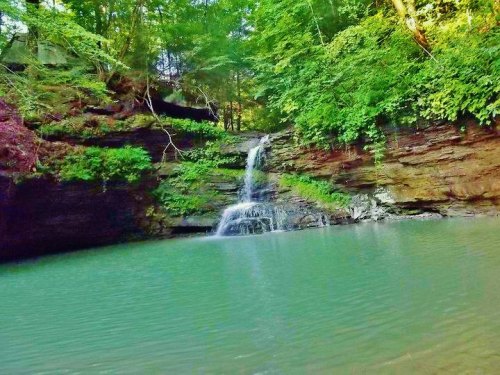 A serene waterfall cascades into a tranquil turquoise pool, surrounded by lush green foliage and rocky cliffs.