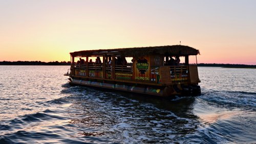 A boat with a thatched roof glides across the water at sunset, creating a serene and picturesque scene.