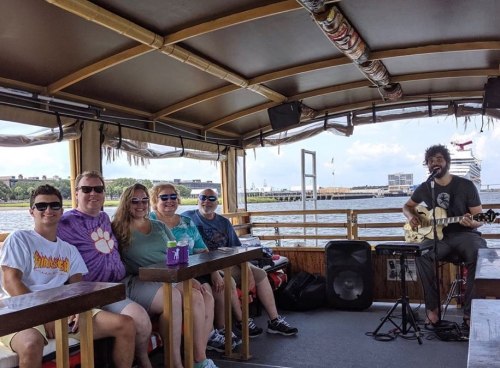 A group of six people sitting on a boat, enjoying music from a guitarist, with a city skyline in the background.