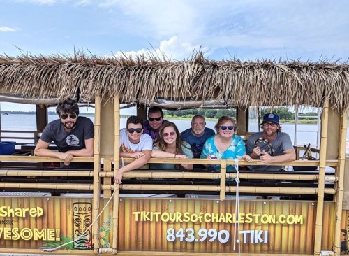 A group of seven people smiles on a tiki boat, surrounded by water and a sunny sky.
