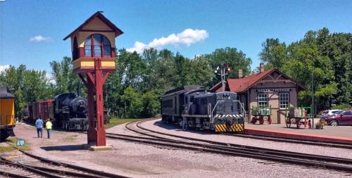A vintage train station with two trains, a signal tower, and green trees under a blue sky.