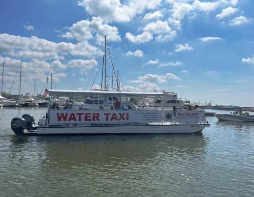 A water taxi with "WATER TAXI" written on the side, docked in a marina with boats and blue skies in the background.