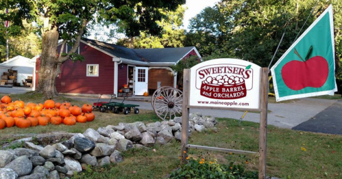 A red barn with pumpkins in front, a sign for Sweetser's Apple Barrel and Orchards, and a green flag with an apple logo.