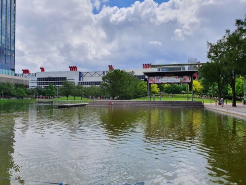 A serene park scene featuring a pond, green trees, and a modern building in the background under a partly cloudy sky.