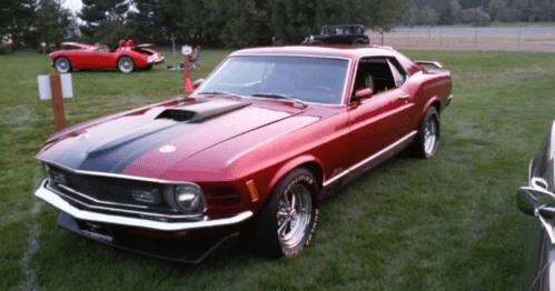 A classic red and black muscle car parked on grass, with vintage cars in the background.
