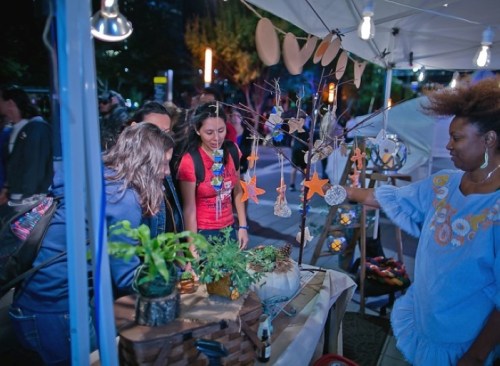 A vendor showcases handmade crafts at a market, while customers admire the unique items on display.
