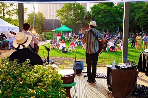 Musicians perform outdoors for an audience seated on the grass, enjoying a sunny day at a community event.