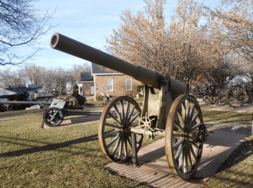 A historic cannon on wheels displayed outdoors, surrounded by trees and other military artifacts.