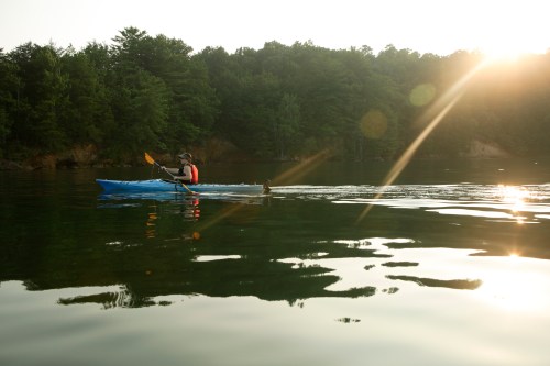 A person kayaking on a calm lake with trees in the background, illuminated by the warm glow of the setting sun.