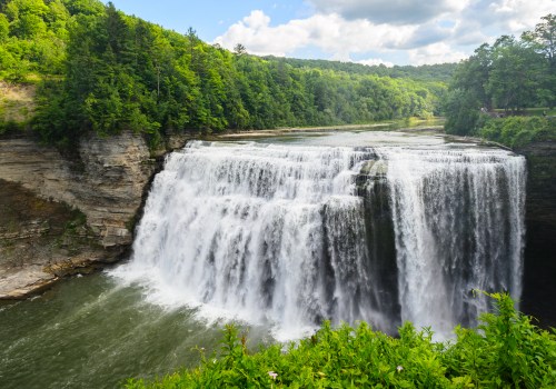 A scenic view of a large waterfall cascading over rocky cliffs, surrounded by lush green trees and a cloudy blue sky.