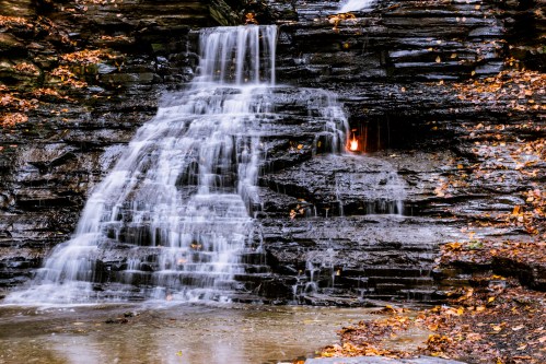 A serene waterfall cascading over dark rocks, surrounded by autumn leaves and a small pool of water below.