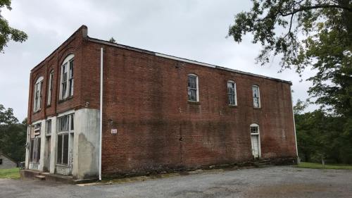 A weathered brick building with boarded windows, set against a cloudy sky and surrounded by greenery.