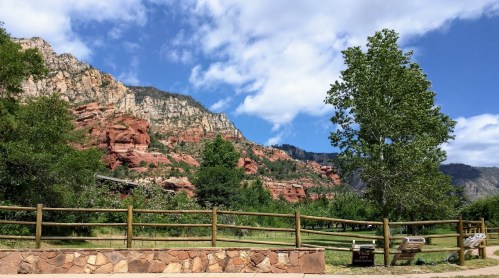 Scenic view of red rock formations and green trees under a blue sky with clouds. A wooden fence in the foreground.
