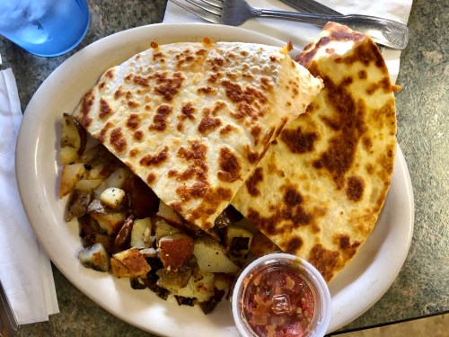 A plate with two large quesadillas, seasoned potatoes, and a small cup of salsa on the side.