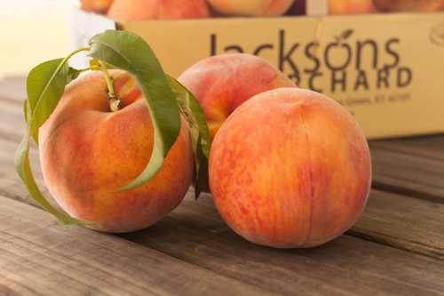 Two ripe peaches with green leaves on a wooden table, with a box of peaches in the background.