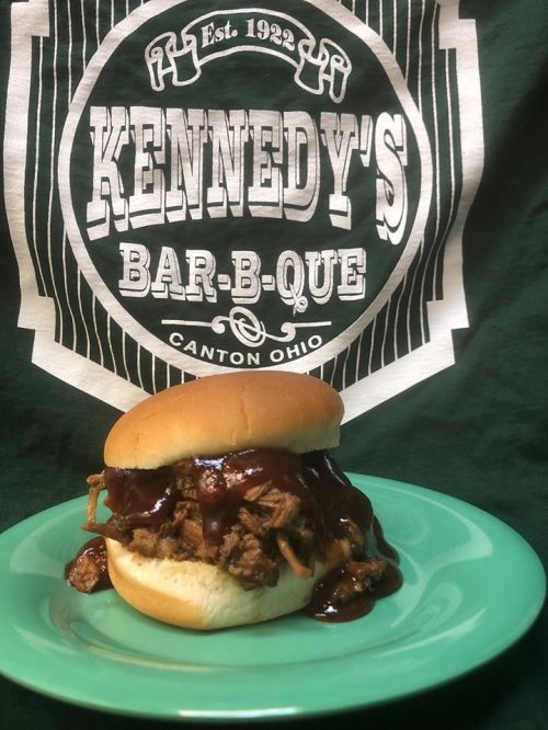 A pulled pork sandwich with barbecue sauce on a green plate, set against a Kennedy's Bar-B-Que shirt background.