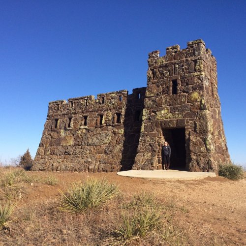 A stone castle-like structure stands against a clear blue sky, with a person standing at the entrance.