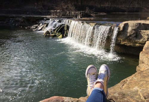 A person’s feet in sneakers resting on a rock by a serene waterfall and calm water.