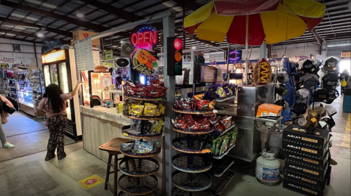 A bustling market stall with snacks, drinks, and an "Open" sign, surrounded by various merchandise and shoppers.