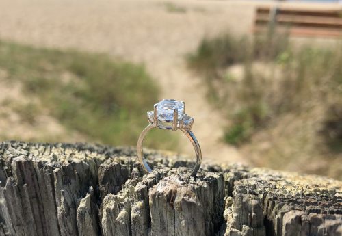 A sparkling engagement ring rests on a weathered wooden post, with a beach and ocean in the background.
