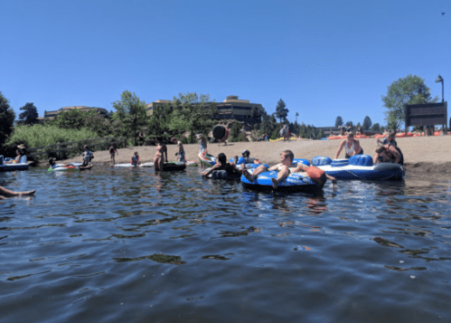 People relaxing on inflatable tubes in a river on a sunny day, with a sandy beach and trees in the background.