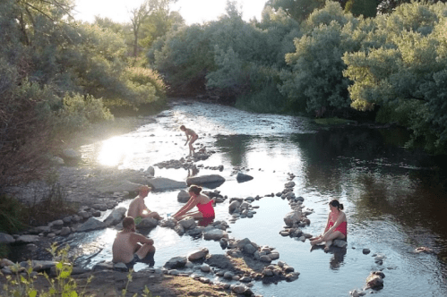 A group of people relaxes by a river, sitting on rocks and enjoying the natural scenery in a sunny setting.