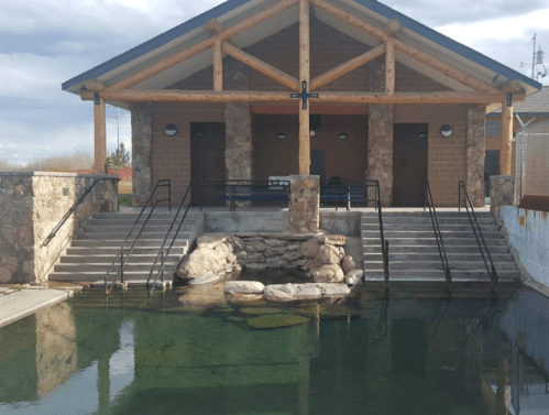 A building with a wooden roof and stone walls, flanked by stairs leading to a clear pond in front.