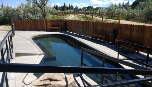 A serene outdoor pool area with a wooden fence, benches, and surrounding greenery under a clear blue sky.