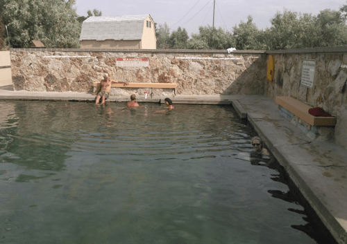 People enjoying a natural hot spring, with a stone wall and a building in the background.