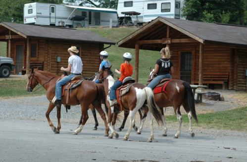 Four people on horseback ride past rustic cabins in a rural setting.