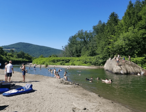 People enjoying a sunny day by a river, swimming and relaxing on rocks, surrounded by lush greenery and mountains.