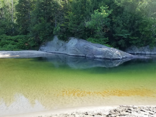 A serene river scene with a rocky bank and lush green trees reflecting in the calm water.