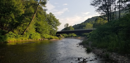 A serene river flows under a bridge, surrounded by lush greenery and hills under a clear sky.