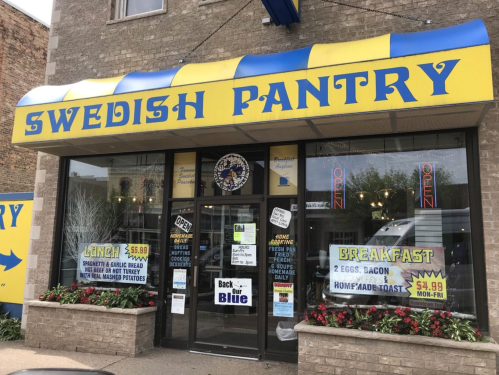 Exterior of Swedish Pantry with a colorful awning, open sign, and flower beds in front. Menu highlights breakfast and lunch.