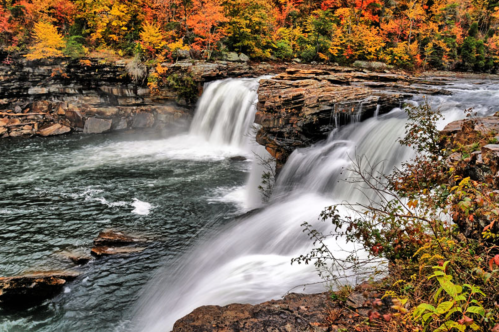 A scenic waterfall cascading over rocks, surrounded by vibrant autumn foliage in shades of orange and yellow.