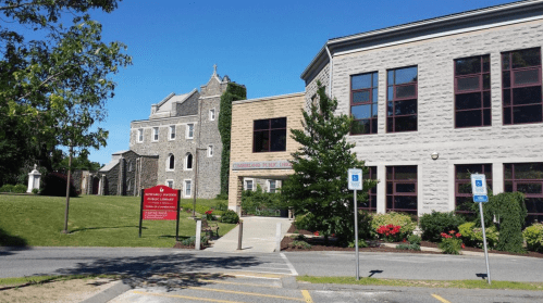 A stone and modern building with greenery, featuring a sign and accessible parking in front. Clear blue sky above.