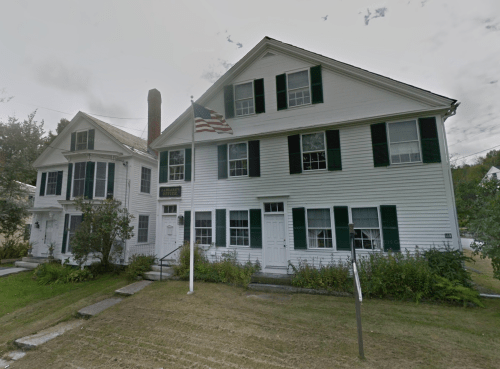 Two white colonial-style houses with green shutters, an American flag, and a grassy lawn.
