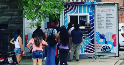 A colorful food truck with a line of people waiting to order, surrounded by greenery and urban buildings.