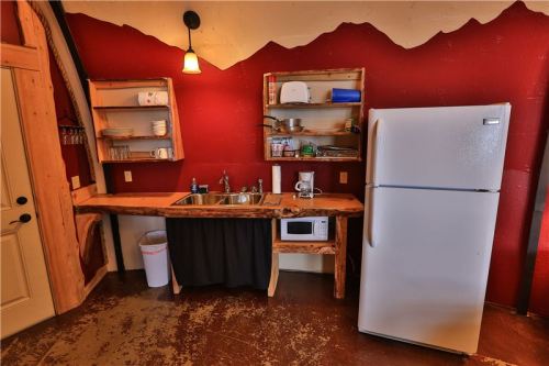 A rustic kitchen with a red wall, wooden shelves, a sink, microwave, and a white refrigerator.