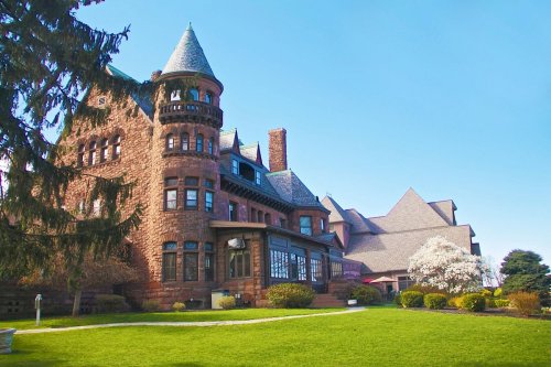 A large, historic stone mansion with a turret, surrounded by green grass and blooming trees under a clear blue sky.