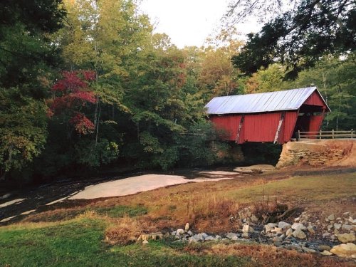 A red covered bridge over a stream, surrounded by trees with autumn foliage in a serene landscape.